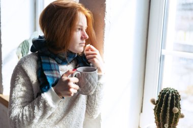 Sıcak eşarp takan ve pencerede duran hasta bir kızın kesilmiş portresi. Kendini soyutlama. Soğuk algınlığı