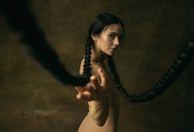 Koyu yeşil arka planda uzun siyah örgülü bej kumaşlı genç balerin portresi.