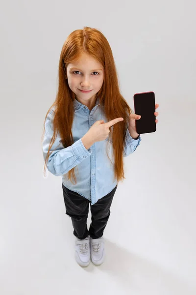 Top view portrait of little girl, child pointing at phone screen isolated over gray background. Social media concept — Stock Photo, Image