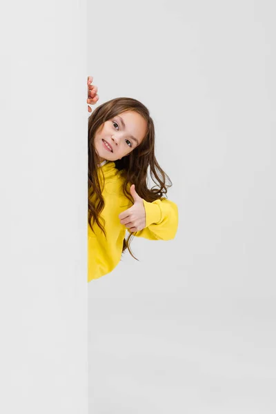 Beskurna porträtt av lekfull liten flicka tittar ut hörnet isolerad över vit bakgrund. Tummen upp — Stockfoto