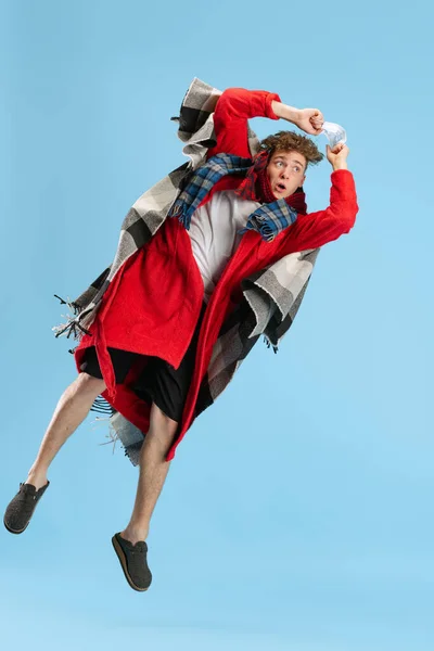 Полнометражный портрет больного мальчика, одетого в клетку, халат и шарф, радостно прыгающего на синем фоне. Восстановление после простуды и гриппа, аллергии — стоковое фото