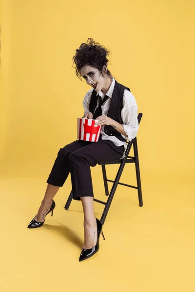 Retrato de cuerpo entero de mujer espeluznante disfrazada de Halloween con canasta de palomitas de maíz sentada en una silla aislada sobre fondo amarillo. Fiesta de Halloween — Foto de Stock