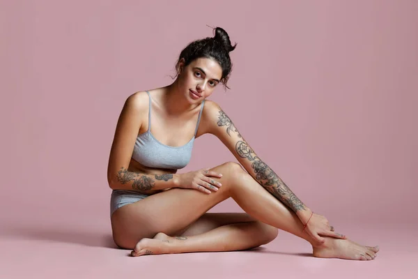 Full-length portrait of beautiful young woman sitting in gray cotton underwear isolated over pink background. — Stock Photo, Image