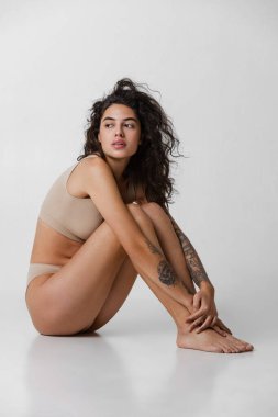 Gri arka planda izole edilmiş bej iç çamaşırlı çekici genç kızın tam boy yan görünüm portresi. Düşünceli bir bakış.