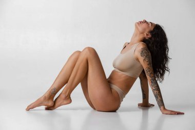 Gri arka planda izole edilmiş bej iç çamaşırlı genç çekici kızın tam boy yatay portresi..