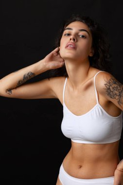 Beyaz pamuklu iç çamaşırlarıyla poz veren, koyu arkaplanda izole edilmiş bir sandalyede oturan güzel kıvırcık bir kadının kırpılmış portresi.