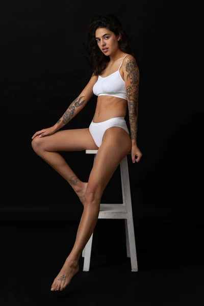 Full-length portrait of beautiful slim girl in white cotton underwear posing on chair isolated over dark background — Stock Photo, Image