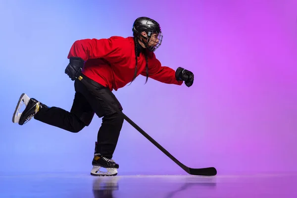 Full-length portrait of professional female hockey player training, dribbling puck isolated over gradient blue purple background.