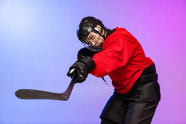 Cropped portrait of professional female hockey player training in special uniform with helmet isolated over gradient blue purple background.