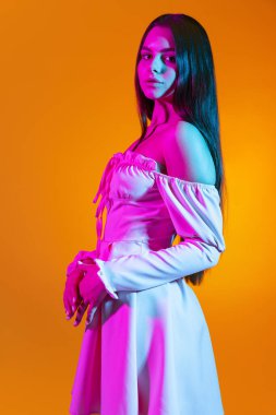 Beyaz elbiseli güzel bir kızın portresi turuncu arka planda izole edilmiş, neon ışıklı bir şekilde poz veriyor.