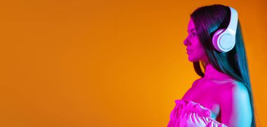 Beyaz kulaklıklı, turuncu arka planda neon ışıkta izole edilmiş müzik dinleyen güzel bir kızın kırpılmış yan görüntüsü. El ilanı