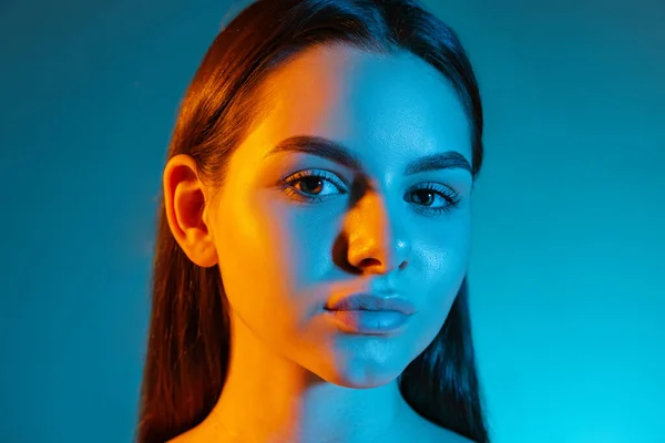 Zbliżenie portret młodej zuchwałej dziewczyny uważnie patrzącej w kamerę, pozującej na niebieskie tło w neonowym świetle — Zdjęcie stockowe