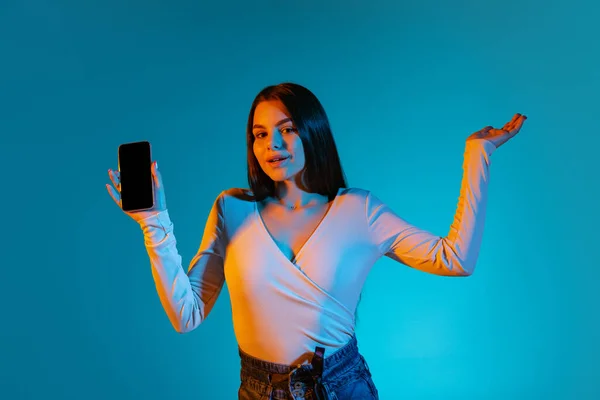 네온 빛을 받은 푸른 배경 위로 핸드폰을 들고 있는 아름다운 소녀의 사진이 찍혀 있다 — 스톡 사진