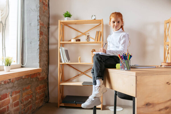 Portrait of little schoolgirl sitting on the table and making notes at home. Doing homework, online studying