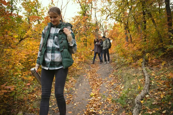 若い女の子は森の中を歩いて、熱いお茶で魔法瓶を保持します。一緒に秋のハイキングをする仲間のグループ — ストック写真