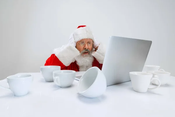 Vágott portré egy idős emberről, aki Mikulás jelmezben ül az asztalnál, tele üres kávéscsészékkel és laptopot néz. — Stock Fotó