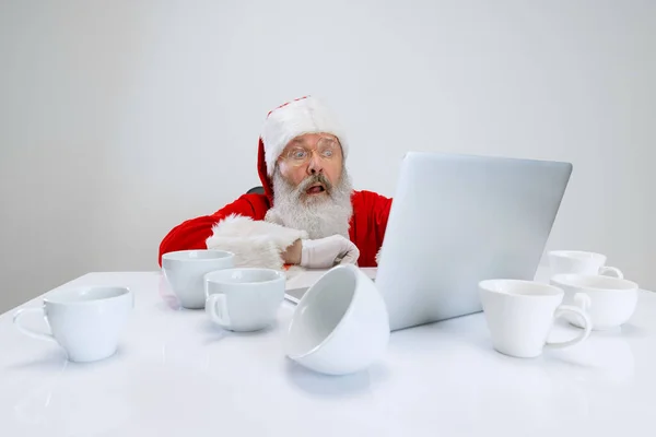 Ausgeschnittenes Porträt eines älteren Mannes im Weihnachtsmannkostüm, der am Schreibtisch voller leerer Kaffeetassen sitzt und auf Laptop schaut — Stockfoto