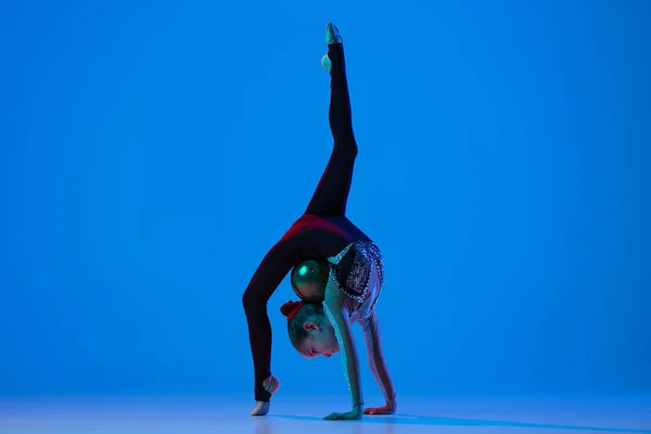 Full-length portret van een klein meisje, professionele ritmische gymnastiek training, het uitvoeren van geïsoleerde over blauwe achtergrond in neon — Stockfoto