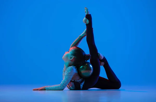 Full-length portret van een klein meisje, professionele ritmische gymnastiek training, het uitvoeren van geïsoleerde over blauwe achtergrond in neon — Stockfoto