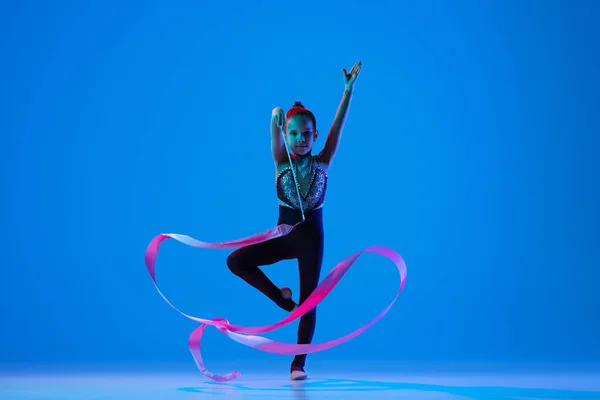 Full-length portret van klein meisje, professionele ritmische gymnastiek training, uitvoeren met roze lint geïsoleerd over blauwe achtergrond in neon — Stockfoto