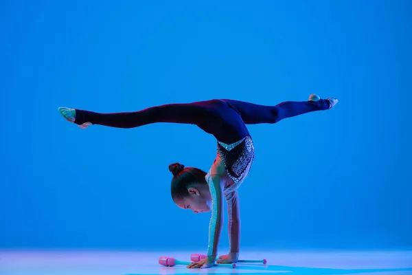 Porträt eines kleinen Mädchens, einer professionellen rhythmischen Turnerin, die auf Bindfäden steht und mit Turnschlägern vor blauem Hintergrund in Neon auftritt — Stockfoto