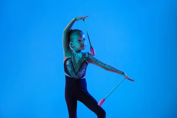 Croppep porträtt av liten flicka, professionell rytmisk gymnast utbildning, utför med gymnastikklubbar isolerade över blå bakgrund i neon — Stockfoto