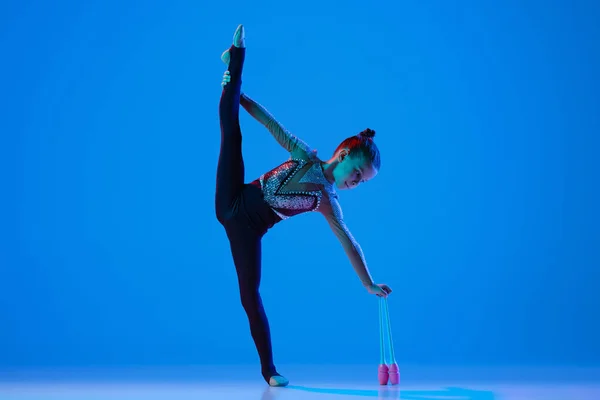 Portret van een klein meisje, professionele ritmische turnster optredend met gymnastiekclubs geïsoleerd over blauwe achtergrond in neon — Stockfoto