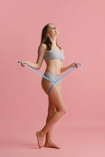 गुलाबी पृष्ठभूमि पर अलग मापने के लिए सेंटीमीटर के साथ ग्रे आंतरिक पोशाक में सुंदर युवा महिला का पूर्ण लंबाई का चित्र . — स्टॉक फ़ोटो, इमेज