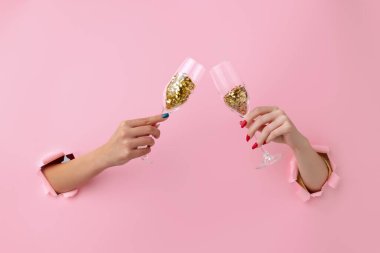Şampanya tutan iki kadın el, konfetili şarap bardağı pembe kağıt arka planda kırılıyor..