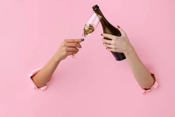 Två kvinnliga händer med flaska vin och champagne glas med konfetti bryter igenom rosa papper bakgrund. — Stockfoto