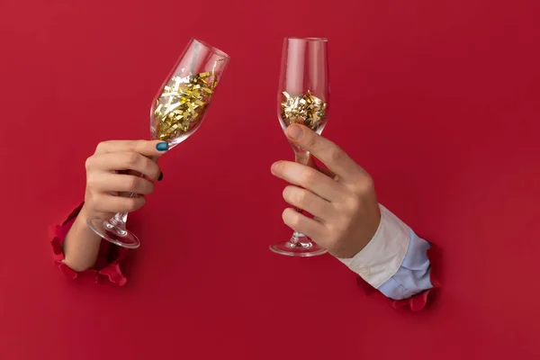 Δύο χέρια, αρσενικό και θηλυκό, κρατώντας φλάουτο σαμπάνιας με κομφετί διαπερνά κόκκινο φόντο χαρτί. Ευτυχισμένο το νέο έτος — Φωτογραφία Αρχείου