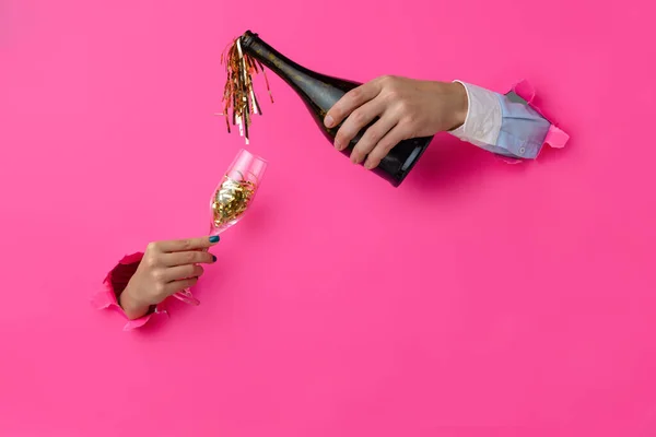 Dos manos rompen el fondo de papel rosa. Hombre vertiendo confeti de botella de vino en flauta de champán — Foto de Stock