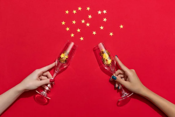 Female hands holding champagne flute with golden confetti isolated over red paper background. Christmas celebration