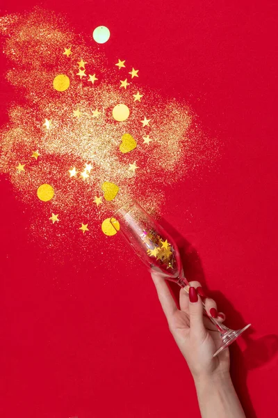 Female hand holding champagne flute with golden confetti isolated over red background