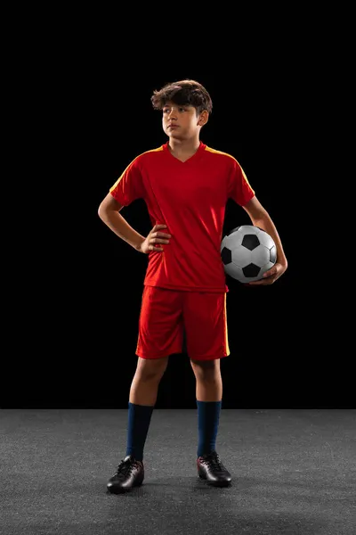 Retrato completo de menino, adolescente, jogador de futebol profissional em uniforme com bola posando isolado sobre fundo preto — Fotografia de Stock