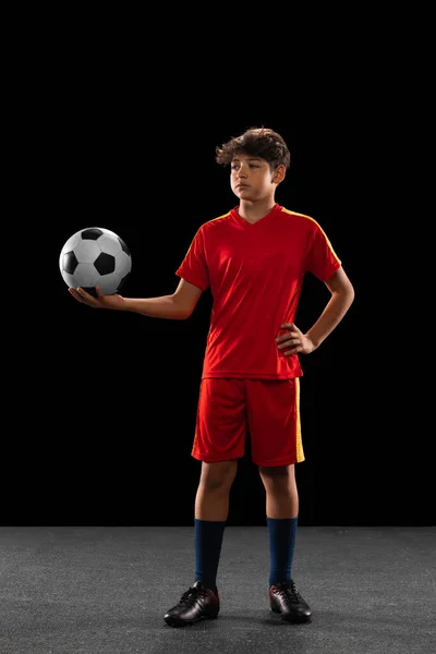 Retrato completo de menino, adolescente, jogador de futebol profissional em uniforme com bola posando isolado sobre fundo preto — Fotografia de Stock
