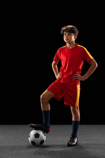 Retrato completo de menino, adolescente, jogador de futebol profissional em uniforme com bola posando isolado sobre fundo preto — Fotografia de Stock