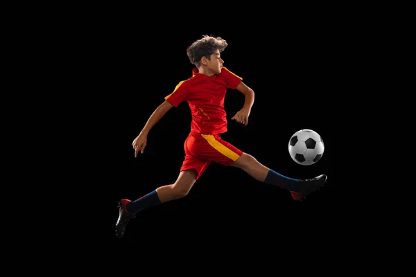 Retrato de niño, adolescente, jugador de fútbol profesional pateando pelota en un salto aislado sobre fondo negro — Foto de Stock