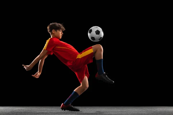 Πορτρέτο του αγοριού, έφηβος, επαγγελματίας ποδοσφαιριστής κλοτσιές μπάλα με γόνατο σε ένα άλμα απομονώνονται σε μαύρο φόντο — Φωτογραφία Αρχείου