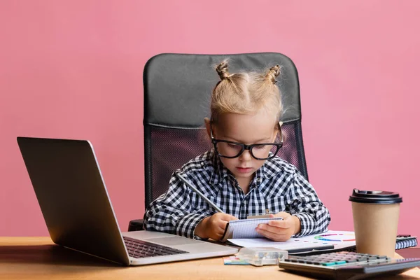 Cropped portrait of cute little girl sitting in front of laptop and making notes isolated over pink background. Schooling — Stock Photo, Image
