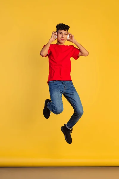 Retrato de cuerpo entero del joven en camiseta roja escuchando música en auriculares y saltando aislado sobre fondo amarillo —  Fotos de Stock