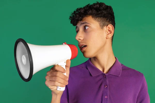 Gesneden portret van jonge jongen in paars T-shirt sprekend in megafoon geïsoleerd over groene achtergrond. Informatieverstrekking — Stockfoto