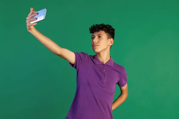 Cropped portrait of young boy in purple T-shirt recordeng taking selfie with phone isolated over green background — Stock Photo, Image