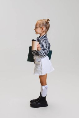 Küçük tatlı bir kızın tam boy yan görünüm portresi, not defteri ve kahve fincanı olan çocuk gri arka planda izole edilmiş sol tarafa bakıyor.