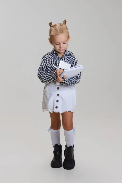 Retrato de comprimento completo de menina bonito, criança em camisa quadriculada fazendo notas isoladas sobre fundo cinza — Fotografia de Stock