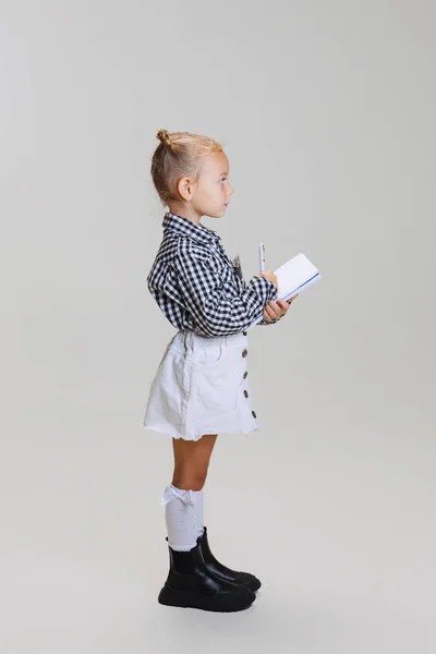 Pełna długość boku widok portret małej uroczej dziewczyny, dziecko w kratkę koszuli z notebooka izolowane na szarym tle — Zdjęcie stockowe