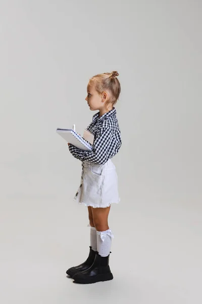 Pełna długość boku widok portret małej uroczej dziewczyny, dziecko w kratkę koszuli z notebooka izolowane na szarym tle — Zdjęcie stockowe