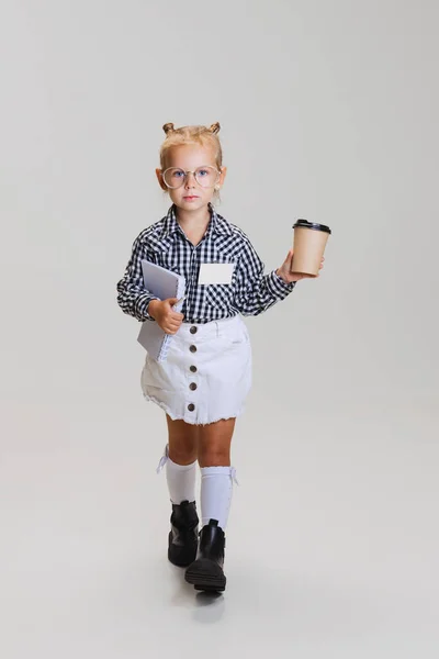 Full-length vista lateral retrato de menina bonito pouco, criança em xadrez camisa andando com notebook e xícara de café isolado sobre fundo cinza — Fotografia de Stock