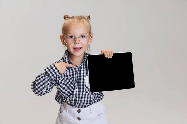 Cropped portrait of cute little girl, child pointing at tablet isolated over gray background. Boss kid — Stock Photo, Image