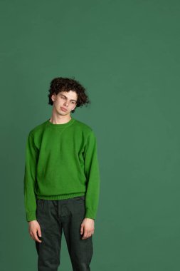 Koyu yeşil arka planda izole edilmiş üzgün, bitkin yüz ifadesi olan yeşil kazaklı genç bir adamın kırpılmış portresi.